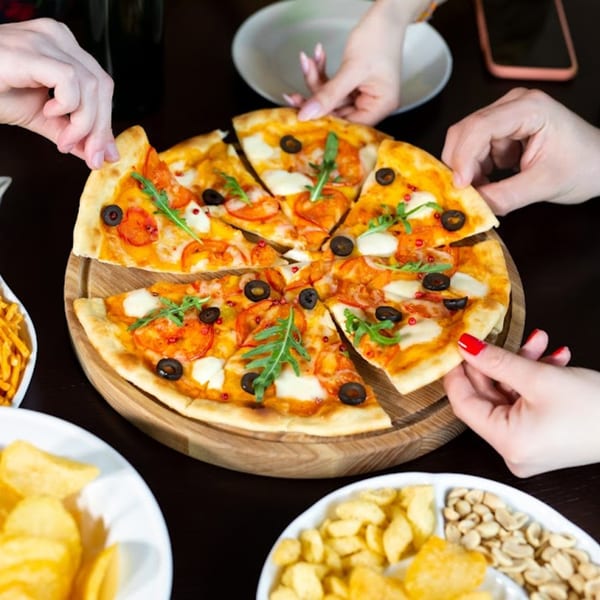 Hands reaching for slices of a Pizzany's Pizzeria pizza with olives, tomatoes, and arugula.